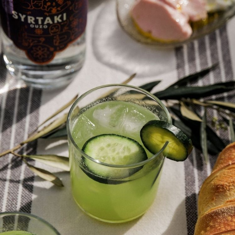 A cucumber cup cocktail in a short glass sitting on a table. A bottle of Syrtaki Ouzo and some snacks can be seen in the background