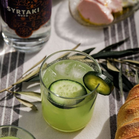A cucumber cup cocktail in a short glass sitting on a table. A bottle of Syrtaki Ouzo and some snacks can be seen in the background