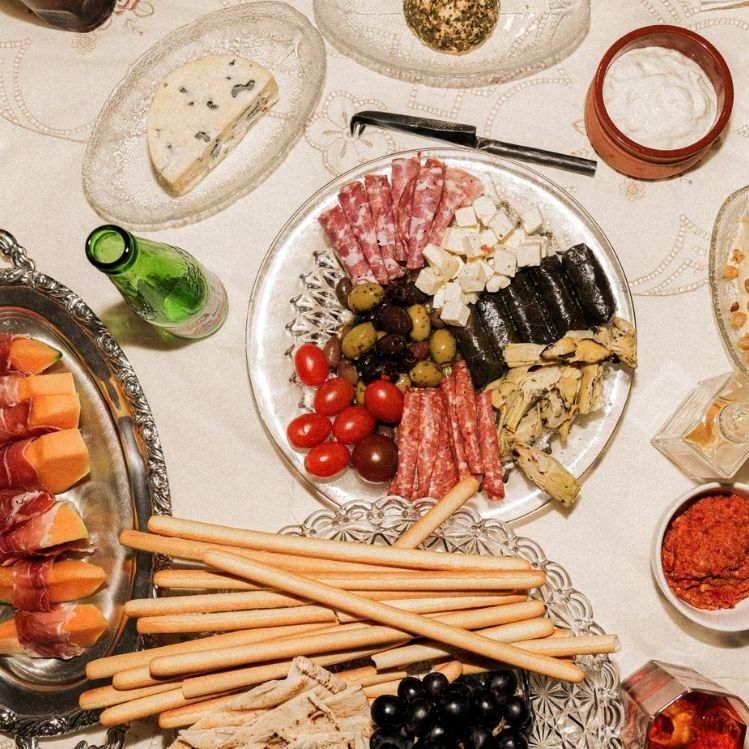 A table laid out with an assortment of meze dishes including cured meats, cheese, fruit, bread and dips. Some drinks can also be seen.