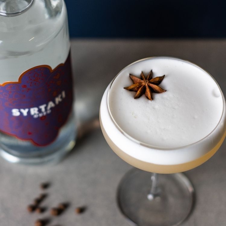 An ouzo sour, pictured from above with a bottle of Syrtaki Ouzo in the background