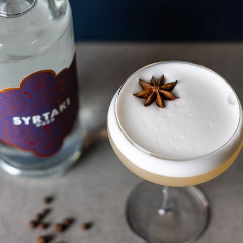 An ouzo sour, pictured from above with a bottle of Syrtaki Ouzo in the background