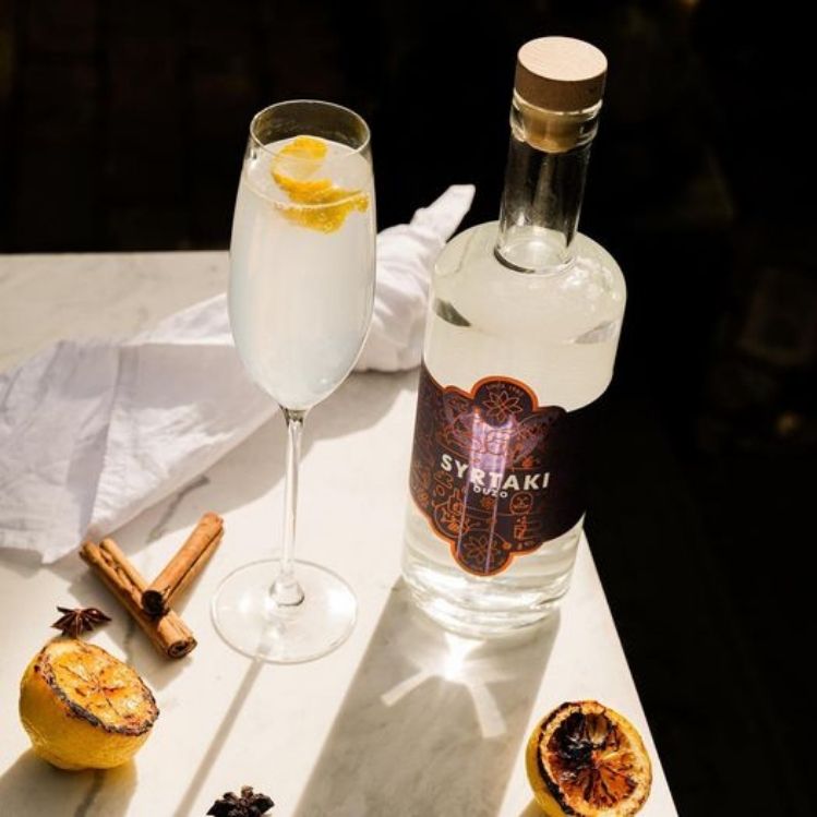 A Greek 75 cocktail in a champagne glass with a bottle of Syrtaki Ouzo some charred lemons and cinnamon sticks scattered around